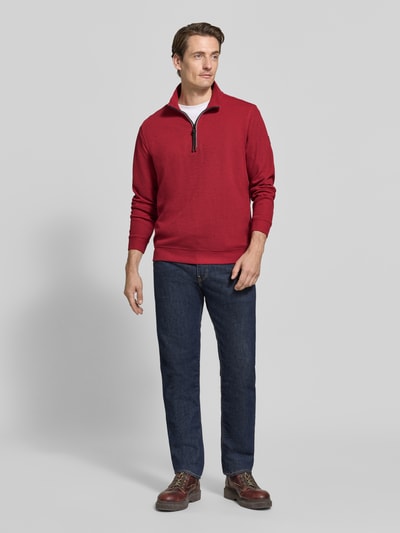 Männermodel trägt Relaxed Fit Sweatshirt aus Baumwoll-Mix Modell 'SELANIK' von LERROS in Rot, kombiniert mit Jeans, Troyer-Kragen, elastische Abschlüsse, Ganzkörperansicht von vorne.