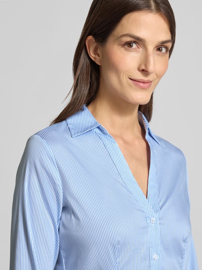 Kobieta modelka nosi Slim Fit Hemdbluse mit V-Ausschnitt od comma, jasnoniebieska w paski, elastyczna bawełna, krój slim fit, długi rękaw, kołnierzyk koszulowy, talia dopasowana, zapięcie na guziki, widok na górną część ciała z przodu.