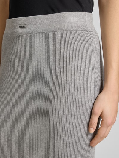 Kobieta nosi Strickrock mit Label-Patch und elastischem Bund von Karo Kauer, mieszanka wiskozy, dopasowany krój, spódnica midi, drobne prążkowanie, naszywka z logo, elastyczny pas, kolor szary. Zbliżenie na dolną część tułowia.