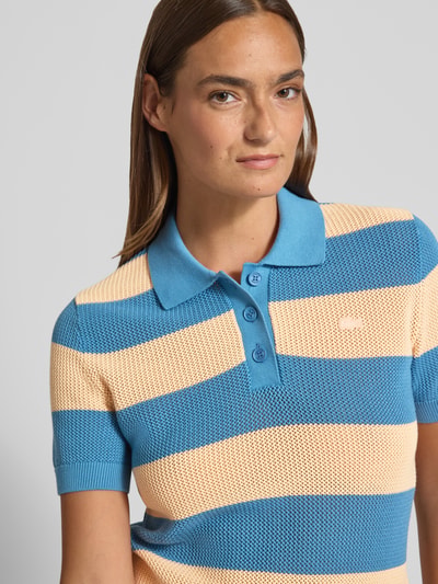 Frauenmodel trägt Regular Fit Poloshirt aus reiner Baumwolle von Lacoste, Optik:Allover-Muster, gestreift, Logo-Patch, Polokragen, V-Ausschnitt, kurze Knopfleiste, Kurzarm, Ärmelabschluss gerippt, Rumpfabschluss gerippt, Materialverarbeitung: Strick, Material: reine Baumwolle, Farbkombination aus Blau und Beige, Nahaufnahme Oberkörper.