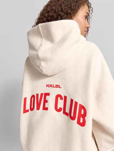 Frauenmodel trägt Hoodie mit Statement-Print Modell 'Love Club' von Karo Kauer aus Reiner Baumwolle im Oversized-Fit, Känguru-Tasche, Kapuze, überschnittene Schultern, beige mit rotem Print, Ganzkörperansicht Rückansicht.