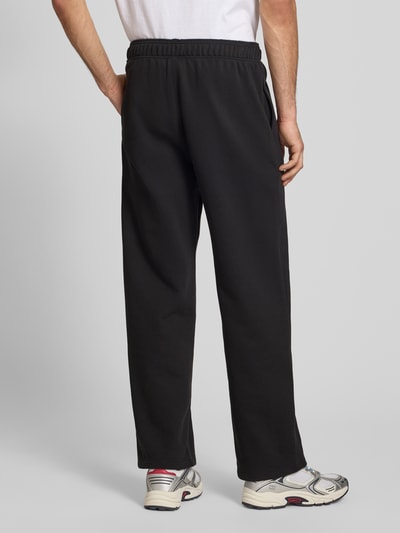 Männermodel trägt schwarze Sweatpants mit Längsnähten und Label-Stitching von CHAMPION aus Baumwoll-Mix, Relaxed-Fit, Ganzkörperansicht Rückansicht.
