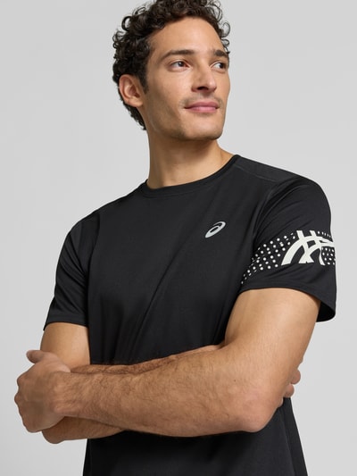 Męski model nosi Regular Fit T-Shirt mit Label-Print Modell 'ICON' von ASICS TIGER, czarny kolor, nadruk z logo, okrągły dekolt, długość tyłu w rozmiarze M: 71 cm, zbliżenie na górną część ciała