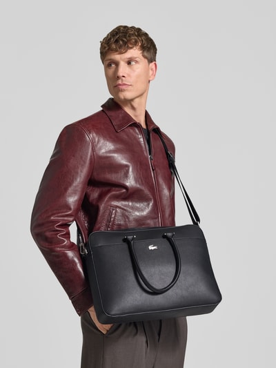 Männermodel trägt Laptoptasche mit Logo-Detail von Lacoste in Schwarz, rote Lederjacke, graue Hose, Ganzkörperansicht von vorne. Kontext: Herren Laptoptasche von Lacoste
