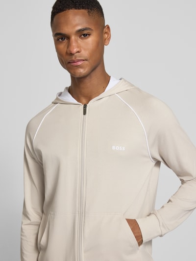 Männermodel trägt Regular Fit Sweatjacke aus Baumwoll-Mix von BOSS in Beige mit weißen Kontraststreifen, Label Stitching, Nahaufnahme Oberkörper.