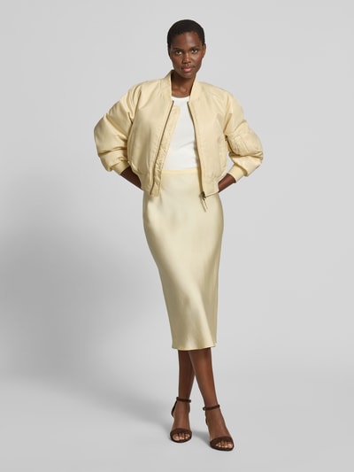 Frauenmodel trägt Bomberjacke mit Stehkragen von Anine Bing in Creme, Regular-Fit, Reißverschluss, Eingrifftaschen, elastische Abschlüsse, kombiniert mit weißem Top und cremefarbenem Rock, Rückenlänge 50 cm, Brustweite 60 cm, Ärmellänge 58 cm, Ganzkörperansicht von vorne.