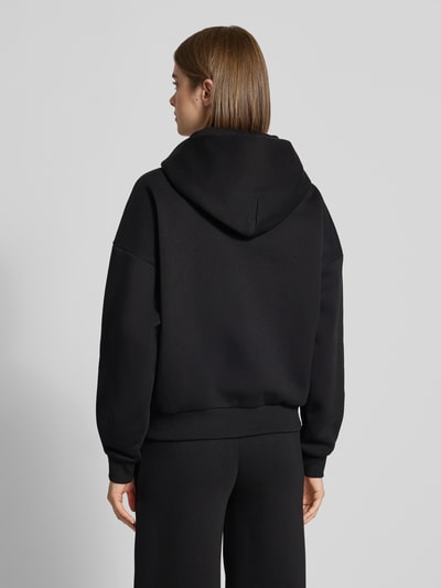 Frauenmodel trägt Relaxed Fit Sweatjacke mit Kapuze von REVIEW in Schwarz, reine Baumwolle, Logo-Print, Känguru-Taschen, Ganzkörperansicht Rückansicht.