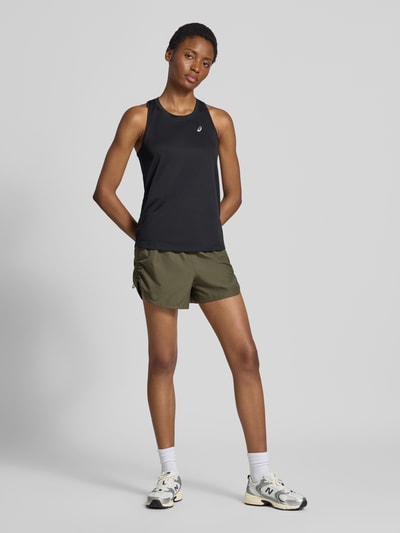 Frauenmodel trägt Shorts mit elastischem Bund Modell 'NAGINO' von ASICS TIGER in Olivgrün mit gerader Passform, reflektierenden Details, rückseitiger Reißverschlusstasche und seitlichen Eingrifftaschen, kombiniert mit schwarzem Top, glatte und matte Textur, Ganzkörperansicht von vorne.