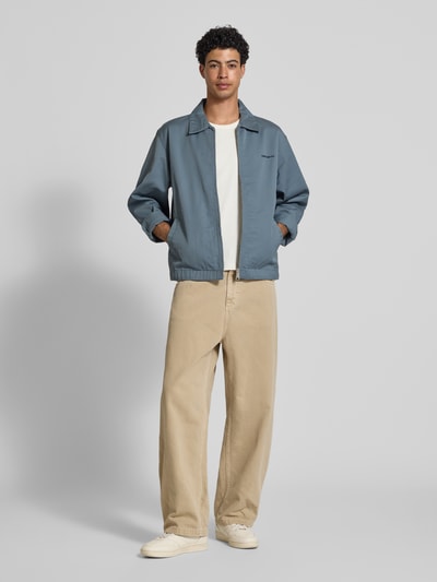 Männermodel trägt Jacke mit Label-Stitching und Eingrifftaschen von Carhartt Work In Progress in Blau, Reißverschluss, beige Hose; Ganzkörperansicht von vorne, Regular Fit, Umlegekragen, strukturierte Oberfläche.