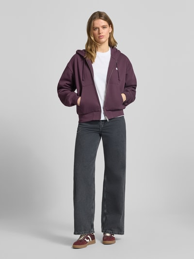 Modelka nosi bluzę rozpinaną z wyhaftowanym logo model 'Casey' od Carhartt Work In Progress z bawełny, krój loose fit, kaptur, zamek błyskawiczny, wpuszczane kieszenie, elastyczne wykończenia, widok całej sylwetki z przodu.