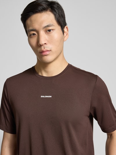 Männermodel trägt braunes Funktionsshirt mit Logo und Rundhalsausschnitt Modell 'Kout' von Salomon, Regular-Fit, Nahaufnahme Oberkörper.