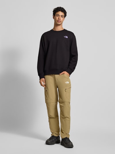 Męski model nosi Bluza z nadrukiem z logo Sweatshirt mit Label-Print od The North Face, bawełna, krój relaxed fit, prążkowane wykończenia, okrągły dekolt, długość tyłu 70 cm, długość rękawów 65 cm, czarna, całkowity widok z przodu.