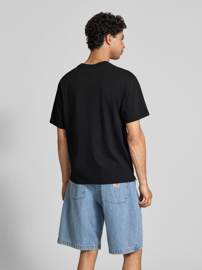 Männermodel trägt T-Shirt mit Label-Stitching Modell 'Madison' von Carhartt Work In Progress in Schwarz, reine Baumwolle, Regular Fit, mittlere Rückenlänge, kombiniert mit blauen Jeansshorts, Ganzkörperansicht Rückansicht.