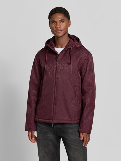 Männermodel trägt Regular Fit Jacke mit Allover-Logo-Print von Emporio Armani in Dunkelrot, Logos und Reißverschluss, Ganzkörperansicht von vorne.