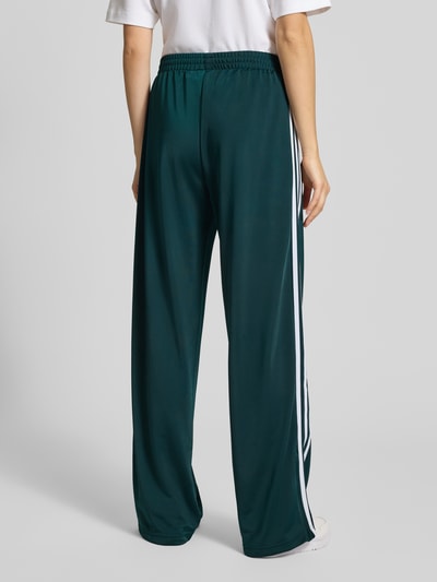 Frauenmodel trägt Sweatpants mit elastischem Bund von adidas Originals in Dunkelgrün mit weißen Streifen, Lockerer-Schnitt, Label-Stitching und Reißverschlusstaschen, Innenbeinlänge bei Größe S: 80 cm, Ganzkörperansicht Rückansicht.