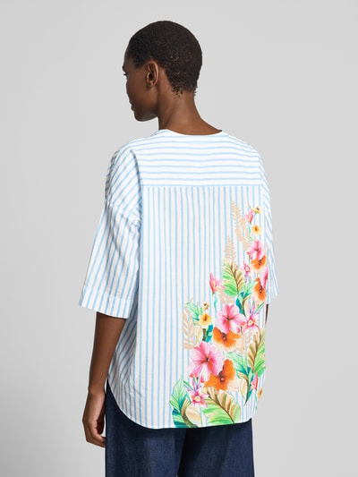 Männermodel trägt Relaxed Fit Bluse mit Motiv-Print von Emily Van den Bergh, bawełna, dekolt w serek, rękaw o dł. 1/2, długość tyłu 48 cm, długość rękawów 24 cm, niebieskie i białe paski, kolorowy nadruk z motywem kwiatów, tekstura gładka, widok całej sylwetki z tyłu.