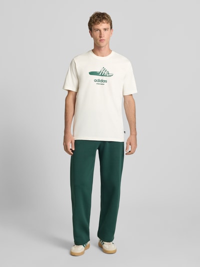 Męski model nosi Regular Fit T-Shirt aus reiner Baumwolle Modell 'SLIDE' od adidas Sportswear, kolor kremowy z zielonym logo, Ganzkörperansicht von vorne.