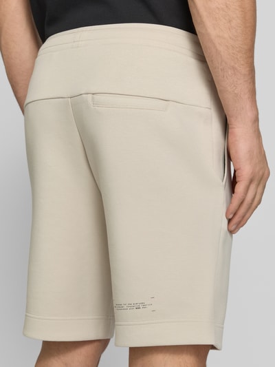 Männermodel trägt Regular Fit Sweatshorts aus Baumwoll-Mix Modell 'JT_GLOBE' von BOSS Green in Optik: Unifarben mit Label-Schriftzug, sichtbaren Nähten, elastischem Bundabschluss, Tunnelzug, seitlichen Eingrifftaschen, seitlicher Reißverschlusstasche, Materialverarbeitung: Sweat, Farbe Beige, Textur weich, Ganzkörperansicht Rückansicht.
