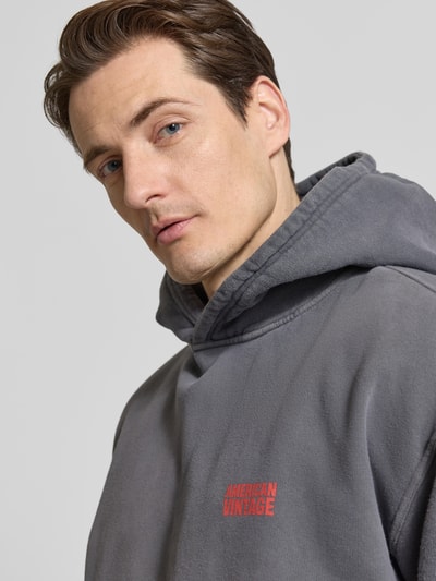 Männermodel trägt Hoodie aus Baumwolle Modell 'PLIZZY' von American Vintage in Dunkelgrau mit Label-Detail, Nahaufnahme Oberkörper. Reine Baumwolle, Lockerer-Schnitt, Kapuze, Känguru-Tasche, Rückenlänge bei Größe M 68 cm, Ärmellänge 60 cm.