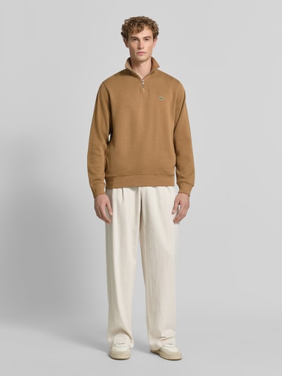 Männermodel trägt Regular Fit Troyer aus reiner Baumwolle von Lacoste in Braun, kombiniert mit heller Hose, Ganzkörperansicht von vorne. Herren Troyer von Lacoste