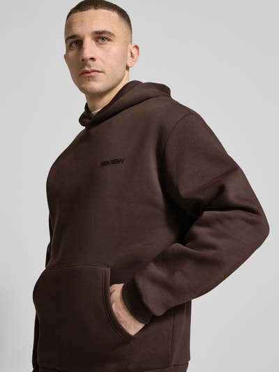 Männermodel trägt Hoodie mit Känguru-Tasche von REVIEW in Dunkelbraun, lockere Passform, Nahaufnahme Oberkörper, Baumwollmischung, Label-Detail.