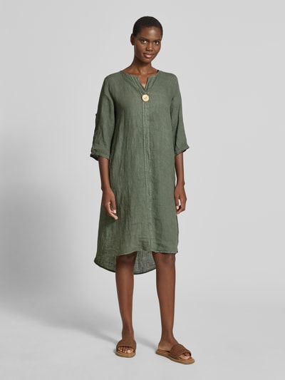 Kobieta modelka nosi knielanges Leinenkleid mit Zierknopf Modell 'Ad44ora' von ZABAIONE, len, linia a, długość do kolan, dekolt w serek, ozdobny guzik, rękaw o dł. 3/4, długość tyłu w rozmiarze S: 107 cm, szerokość w rozmiarze S: 47 cm, długość rękawów w rozmiarze S: 30 cm, całość w oliwkowej zieleni, gładka tekstura, widok całej sylwetki z przodu