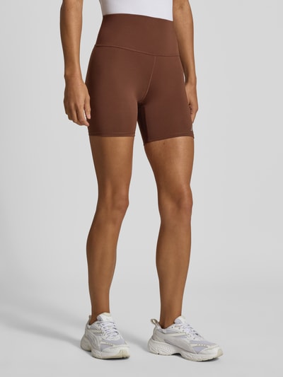 Frauenmodel trägt High Rise Radlershorts mit Logo-Print Modell 'HARMONY' von New Balance in Braun, körpernaher Schnitt, Nahaufnahme Oberkörper.