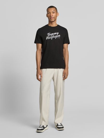 Männermodel trägt Regular Fit T-Shirt aus reiner Baumwolle von Tommy Hilfiger in Schwarz, unifarben mit Label Print, Rundhalsausschnitt, kurze Ärmel, Jersey-Material, Ganzkörperansicht von vorne.