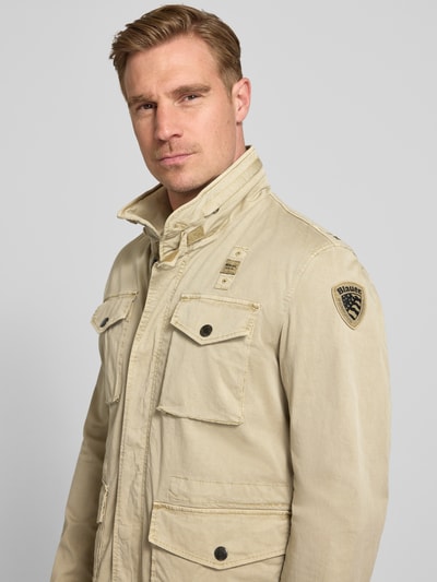 Męski model trägt kurtkę Jacke mit Stehkragen und Pattentaschen Modell 'Ashmont' von Blauer Usa, mieszanka bawełny i elastanu, krój regular fit, jasny beż, kieszenie z patką, stójka, naszywka na ramieniu, Ganzkörperansicht von vorne
