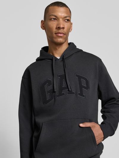 Männermodel trägt 'Hoodie mit Logo-Detail und Känguru-Tasche' von GAP in Schwarz, Baumwoll-Mix, loose fit, Nahaufnahme Oberkörper.