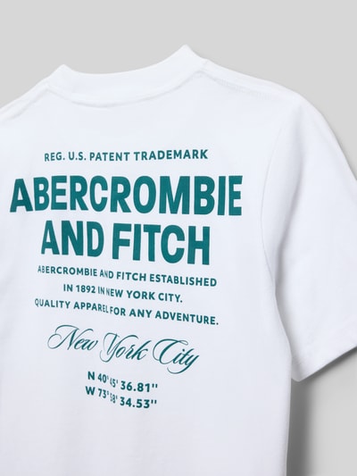 Männermodel trägt T-Shirt aus Baumwoll-Mix mit Label-Print Modell 'CITY STACK' von Abercrombie&Fitch, weiß mit grünem Print, Detailaufnahme Rücken, Kinder T-Shirt, gerade Passform, gerippter Rundhalsausschnitt, Rückenlänge bei Größe 134: 55 cm.