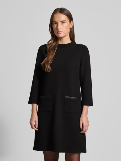 Frauenmodel trägt Minikleid mit 3/4-Ärmel von More & More aus schwarzem Material-Mix mit Baumwoll-Anteil, gerippter Rundhalsausschnitt, taillierter Schnitt, Eingrifftaschen, glatte Textur, Nahaufnahme Oberkörper.
