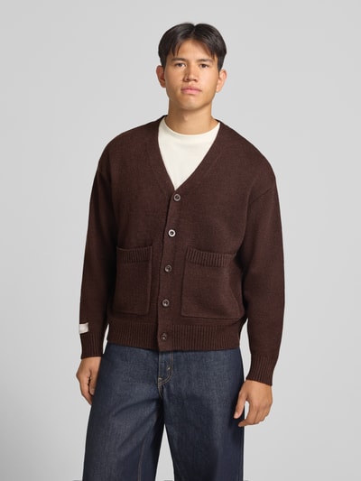 Männermodel trägt Cardigan mit rückseitigem Motiv und V-Ausschnitt von ANOTHER COTTON LAB, braun, gerade Form, Knopfleiste, Ganzkörperansicht von vorne. Rückenlänge 62 cm, Ärmellänge 57 cm, aufgesetzte Taschen, Label-Patch.