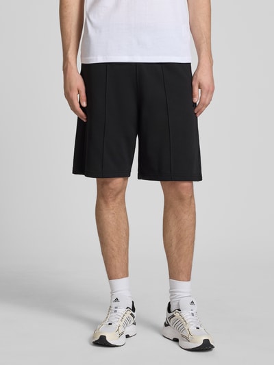 Männermodel trägt Bermudas mit elastischem Bund und Label-Stitching von CHAMPION aus Baumwoll-Mix, Schwarz, Regular-Fit, Längsnähte, Ganzkörperansicht von vorne.