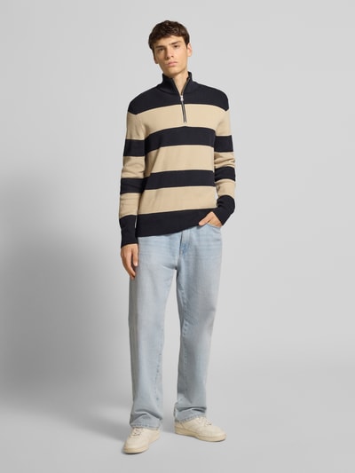 Männermodel trägt Regular Fit Troyer aus Baumwoll-Mix Modell 'PHIL' von Only & Sons mit gestreiftem Allover-Muster in Beige und Dunkelblau, Feinstrick mit Stehkragen und halbem Reißverschluss, Ganzkörperansicht von vorne.