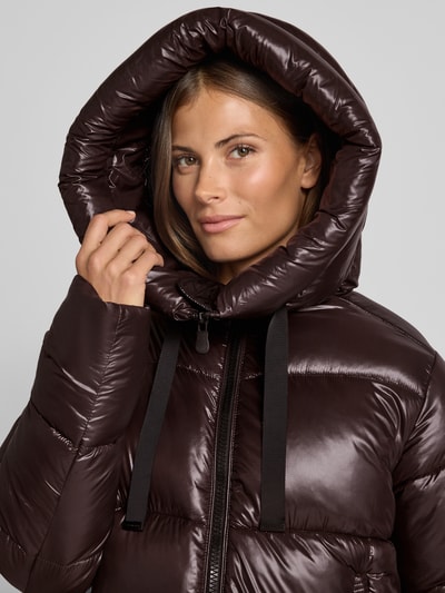 Frauenmodel trägt Steppmantel mit Kapuze und Label-Patch Modell 'ISABEL' von Save The Duck, glänzende braune Oberfläche, Nahaufnahme Oberkörper. Gerade Passform, Tunnelzug an der Kapuze, sichtbarer Zweiwege-Reißverschluss, Kontext: Damen Steppmantel, seitliche Reißverschlusstaschen, Maße bei Größe S: Rückenlänge 90 cm, Schulterbreite 45 cm, Brustweite 54 cm, Ärmellänge 64 cm.