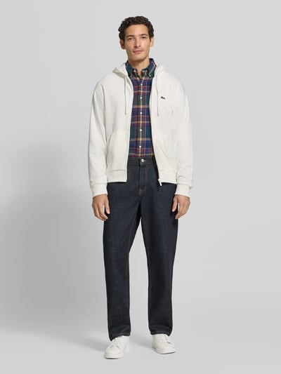 Männermodel trägt Regular Fit Sweatjacke aus Baumwoll-Mix von Lacoste in Weiß zu Jeans und Hemd, Ganzkörperansicht von vorne. Herren Sweatjacke von Lacoste.