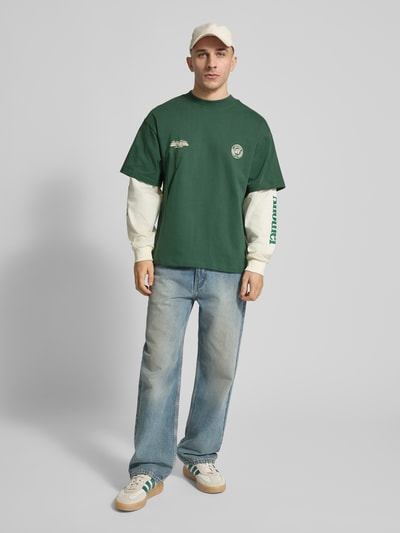 Männermodel trägt Relaxed Fit Longsleeve im Double-Layer-Look Modell 'Sunday Miles' von ANOTHER COTTON LAB in Grün und Creme, Jeans, Sneaker, Statement- und Label-Prints, reine Baumwolle, Ganzkörperansicht von vorne.