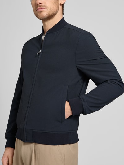 Męski model nosi Bomberjacke mit Eingrifftaschen Modell 'Indro' od JOOP! Collection, materiał z dodatkiem stretchu, proste cięcie, kolor ciemnogranatowy, widoczne kieszenie boczne, krótkie ściągacze, zamek błyskawiczny, gładka tekstura, widok od frontu, zbliżenie na górną część ciała.