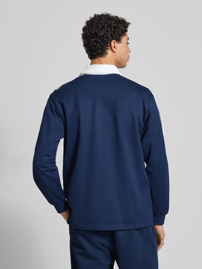 Männermodel trägt Poloshirt mit Label-Stitching von adidas Originals in Dunkelblau mit weißen Details, Rückenlänge 73 cm, Ganzkörperansicht Rückansicht.