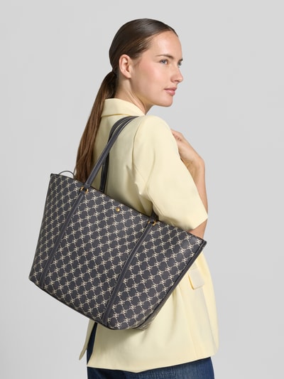 Frauenmodel trägt Tote Bag mit Magnetverschluss Modell 'PORTA' von MANGO mit Allover-Label-Print, schmale Schulterriemen, Ganzkörperansicht von vorne.