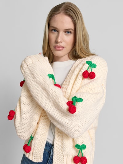 Frauenmodel trägt Oversized Strickjacke mit Zier-Applikationen von Apricot in Creme mit roten und grünen Details, Grobstrick, Nahaufnahme Oberkörper.