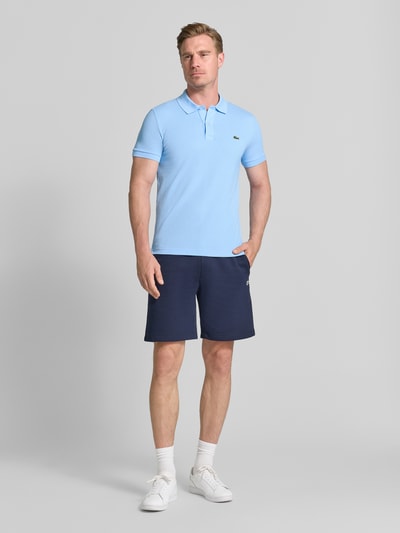 Männermodel trägt Slim Fit Poloshirt aus reiner katoen van Lacoste in lichtblauw, gecombineerd met donkerblauwe shorts; ganzkörperansicht von vorne.