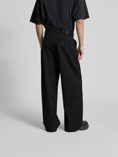 Męski model nosi Straight Fit Hose aus Schurwoll-Mix von VETEMENTS, czarny, elastyczny materiał, kieszenie z tyłu, kanty, krój straight fit, zapięcie na hak i zamek, długość nogawki wewnętrznej w rozmiarze M 77 cm, szerokość w rozmiarze M 44 cm, całkowity widok z tyłu.
