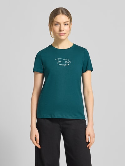 Modelka kobieca nosi Regular Fit T-Shirt aus Bio-Baumwoll-Mix od Tom Tailor, krój regular fit, jednolity kolor, okrągły dekolt, krótki rękaw, aplikacja z napisem, materiał bawełna organiczna-mix, widok całej sylwetki z przodu.