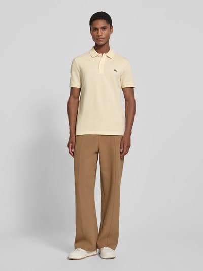 Männermodel trägt Slim Fit Poloshirt aus reiner Baumwolle von Lacoste in Beige, kombiniert mit brauner Hose, Ganzkörperansicht von vorne.