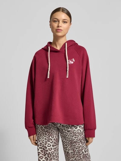 Frauenmodel trägt Relaxed Fit Hoodie mit Motiv-Print von Marc Cain, bordeauxrot, elastischer Baumwoll-Mix, Kapuze, Statement-Print, Ganzkörperansicht von vorne.