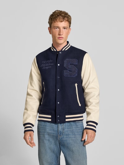 Männermodel trägt College-Jacke aus Woll-Mix mit Eingrifftaschen von Superdry in Blau und Creme, Regular Fit, Ganzkörperansicht von vorne.