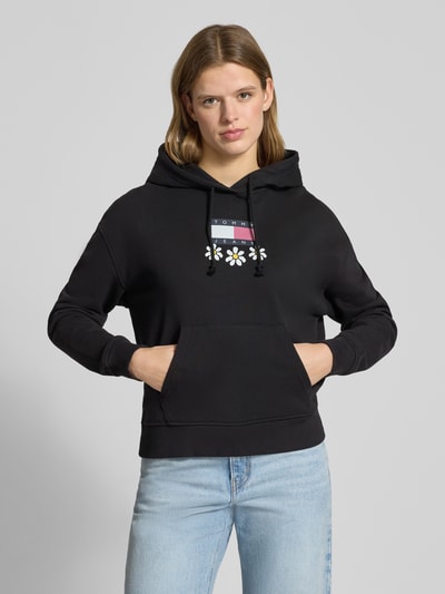 Modelka nosi czarny Hoodie mit Label- und Motiv-Print Modell 'DAISY FLAG' von Tommy Jeans z bawełnianej mieszanki, luźny krój, gładka faktura, z kapturem i kieszenią typu kangurka, widok na cały tułów z przodu.