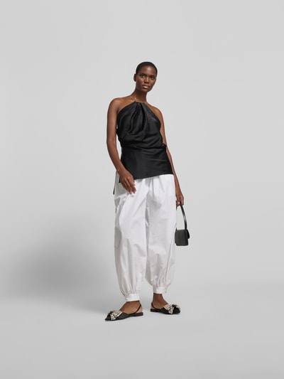 Frauenmodel trägt Top aus Satin mit Viskose-Anteil von Victoria Beckham in Schwarz, körpernahe Passform, Neckholder, ärmel- und schulterlos, Material-Mix, kombiniert mit weißer Hose, glatte Textur, Ganzkörperansicht von vorne.